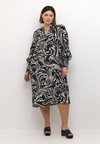 Robe noire et blanche avec un motif marbré tourbillonnant, à manches longues et décolleté en V. Associée à des sandales à plateforme noires. Tissu lisse.
