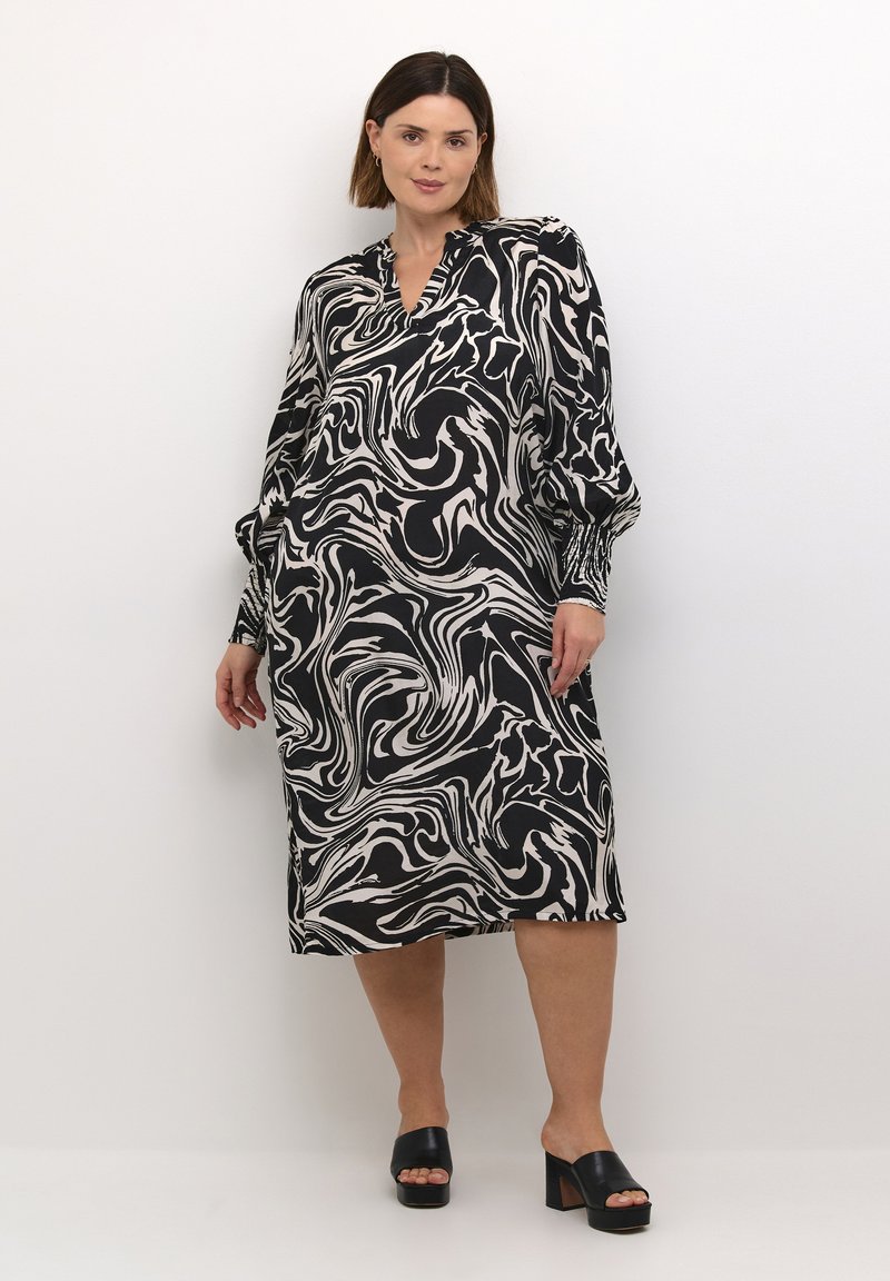 Robe noire et blanche avec un motif marbré tourbillonnant, à manches longues et décolleté en V. Associée à des sandales à plateforme noires. Tissu lisse.