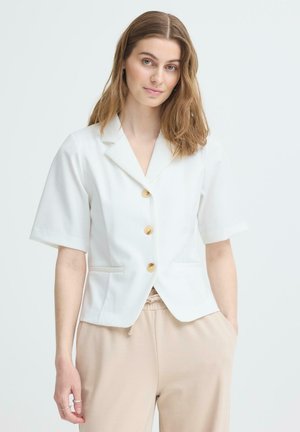 Jeune femme aux cheveux châtain clair portant un blazer blanc à manches courtes boutonné et un pantalon beige à cordon, posant devant un fond uni.