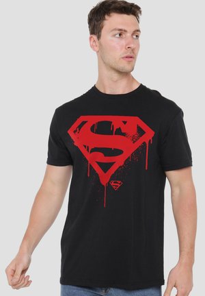 Young man wearing black t-shirt with large red dripping Superman logo, looking to his left against a plain background.