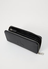 Portefeuille en cuir noir avec logo "CK" en relief, disposant d'une fermeture éclair argentée, de multiples compartiments et d'un design épuré.