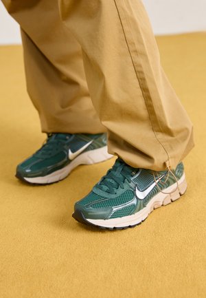 Osoba nosząca beżowe spodnie i zielone buty sportowe Nike stoi na żółtej podłodze, a mankiety spodni są ściągnięte regulowanymi sznurkami.