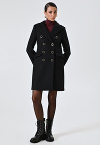 Cappotto doppiopetto nero con dettagli in metallo dorato, tessuto strutturato e tasche laterali. Abbinato a stivali neri stringati e collant scuri.