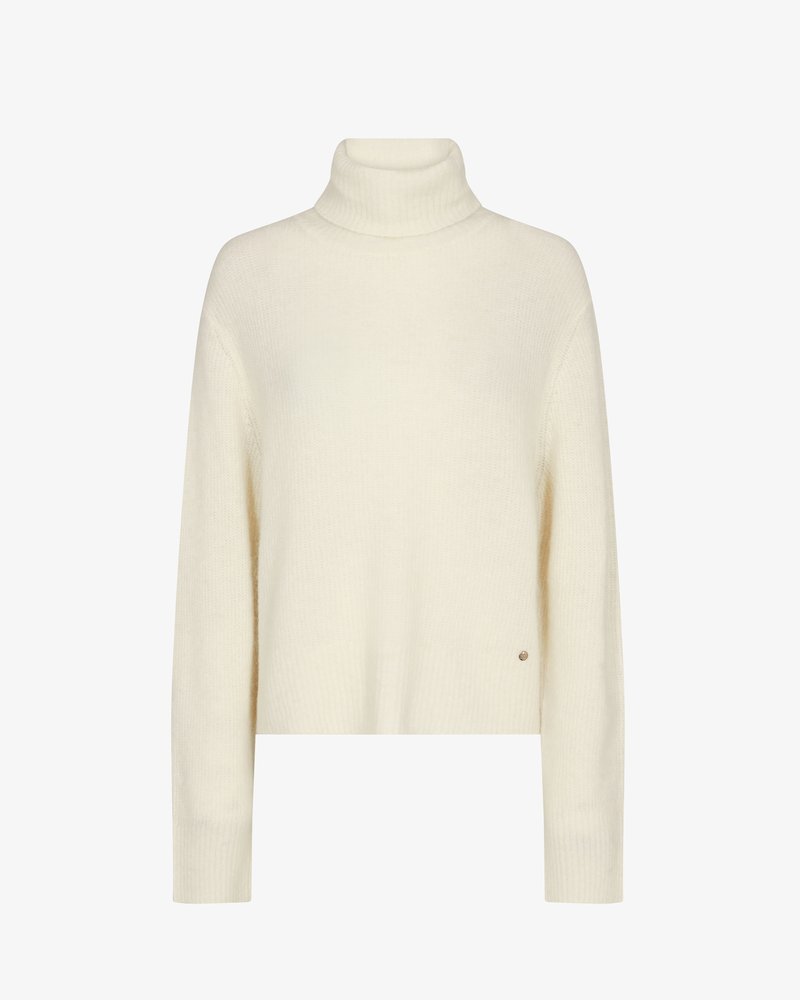 Cream-colored knit turtleneck sweater with long sleeves and ribbed cuffs, showing a small round metal logo near the hem.