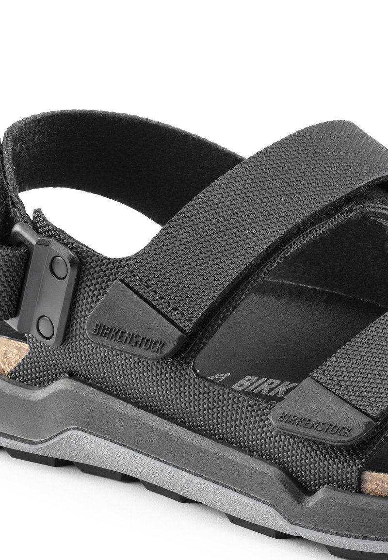 Sandales noires avec des brides synthétiques texturées, des boucles réglables et une semelle en liège avec une semelle en caoutchouc gris offrant un bon soutien.