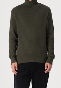 Pull à col roulé côtelé vert foncé avec une finition texturée, manches longues et coupe ajustée. Associé à un pantalon noir.