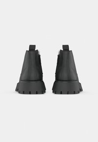 Paire de bottines noires à semelles épaisses et languettes, vues de dos sur un fond clair uni.