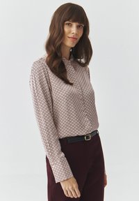 Langärmlige Bluse in Hellrosa mit geometrischem Muster in Dunkelburgunder. Sie hat einen Hemdkragen und wird mit dunklen Hosen kombiniert.