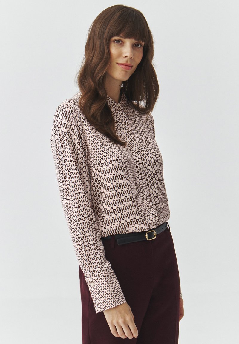 Langärmlige Bluse in Hellrosa mit geometrischem Muster in Dunkelburgunder. Sie hat einen Hemdkragen und wird mit dunklen Hosen kombiniert.