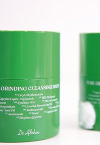 Zielony okrągły pojemnik oznaczony "Balsam do czyszczenia do mielenia" z białym napisem; gładka tekstura; minimalistyczny design; obok częściowo widoczny inny pojemnik.