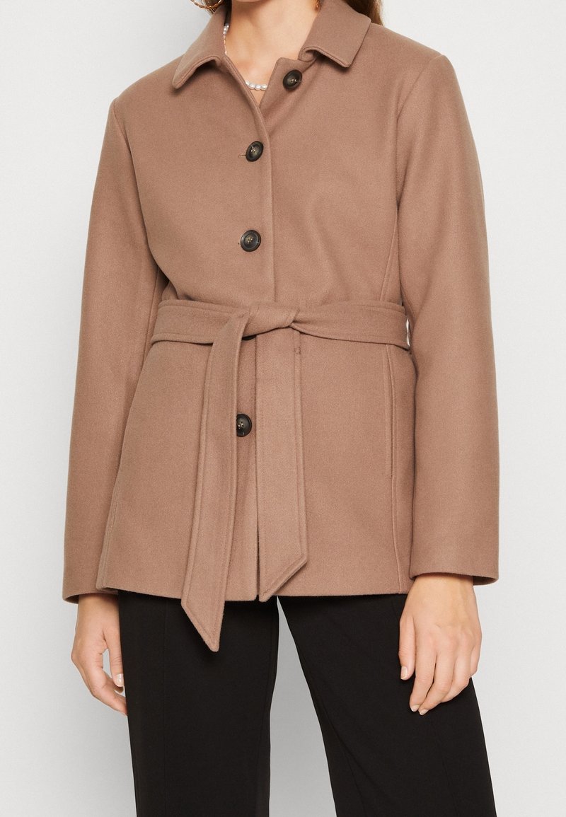 Femme portant un manteau ceinturé couleur camel avec quatre boutons foncés, assorti à un pantalon noir sur un fond uni.