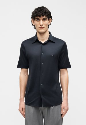 Mężczyzna w czarnej, krótkim rękawem, koszuli na guziki i szarych spodniach, stojący na tle jednolitego, jasnego tła, zwrócony przodem.