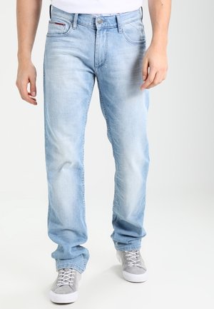 Man wearing light blue jeans and gray sneakers, standing against a plain white background.