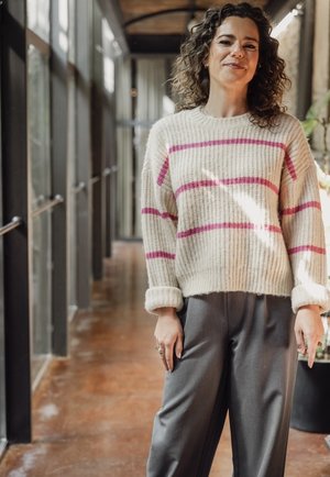 Stevige crèmekleurige trui met horizontale roze strepen, geribbelde textuur en opgerolde manchetten, gecombineerd met losse grijze broek.
