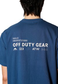 Blå bomullströja med korta ärmar. Bakre delen har vit tryckt text: "HALO 196819721981 OFF DUTY GEAR 223 ATW B-583" och en bergslogo.