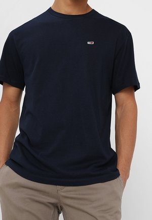 Mies, joka pukeutunut tummansiniseen lyhythihaiseen pyöreäkauluksiseen t-paitaan, jossa on pieni suorakulmainen logo rinnassa, ja beigenvärisiin housuihin, kädet taskuissa.