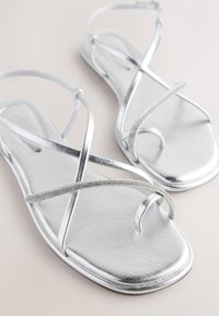 Sandale plate argintii cu barete încrucișate, cu finisaj metalic și decorate cu un mic accent de stras. Textură moale și netedă.