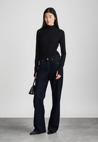 Μαύρη μπλούζα με μακριά μανίκια και ψηλό λαιμό, σε συνδυασμό με σκούρα μπλε παντελόνια με φαρδιά γραμμή. Το μοντέλο κρατά μια μικρή μαύρη τσάντα και στέκεται σε ουδέτερο φόντο.