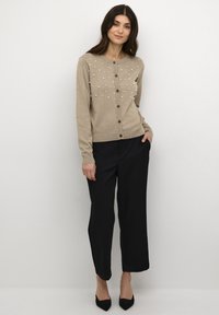 Bēša kardigāns ar pērļu rotājumiem, apaļa izgriezuma, ar pogām priekšā, ribotas aproces un apakšmalu. Kombinēts ar brīvām melnām biksēm un augstpmaka augstiem apaviem.