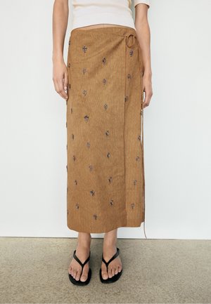 Personne portant une jupe midi portefeuille marron avec de petits motifs brodés foncés et des sandales noires à brides, debout devant un fond uni.