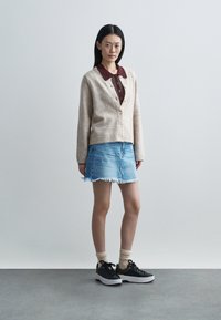 Femme debout portant un cardigan beige boutonné sur une chemise bordeaux à col, une jupe en jean effilochée, des chaussettes beiges et des baskets noires sur un sol gris.
