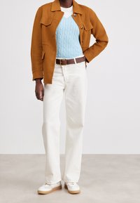 Osoba ubrana w rdzawą kurtkę na jasnoniebieski sweter w warkocze, białe spodnie z brązowym paskiem i białe sneakersy.