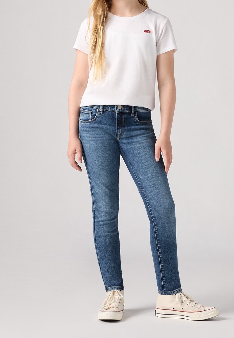 Vit t-shirt med kort ärm och röd logotyp, kombinerad med blå skinny jeans och beige canvas sneakers med röda accenter.