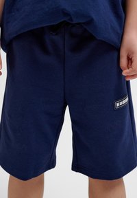 Marina blå bomullsshorts med elastisk midja och liten logotyp på högra sidan. Slät textur, knälång design.