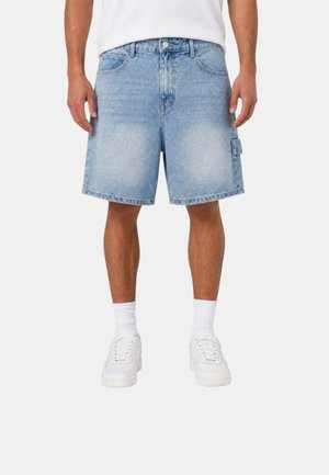 Uomo che indossa pantaloncini di jeans azzurri, calzini bianchi e scarpe da ginnastica bianche, in piedi davanti a uno sfondo chiaro uniforme.