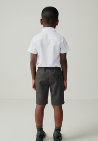 Weißes kurzärmeliges Hemd mit strukturiertem Kragen; graue Shorts mit einer Gesäßtasche; kombiniert mit schwarzen Schuhen und gestreiften Socken.