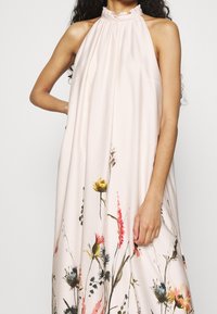 Mujer vestida con un vestido sin mangas de corte halter en un suave rosa pálido, con un estampado floral que presenta flores amarillas, rosas y azules cerca del dobladillo.