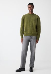 Zaļš teksturēts džemperis ar apaļu kakla izgriezumu, kopā ar gaiši pelēniem taisnajiem džinsiem un melnām kurpēm, ko nēsā stāvoša modeļa figūra.