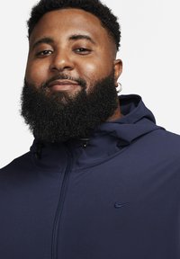 Mörkblå Nike-huvjacka med en slät textur, utrustad med hel dragkedja och justerbara dragsnören vid huvan. Liten logotyp på bröstet.