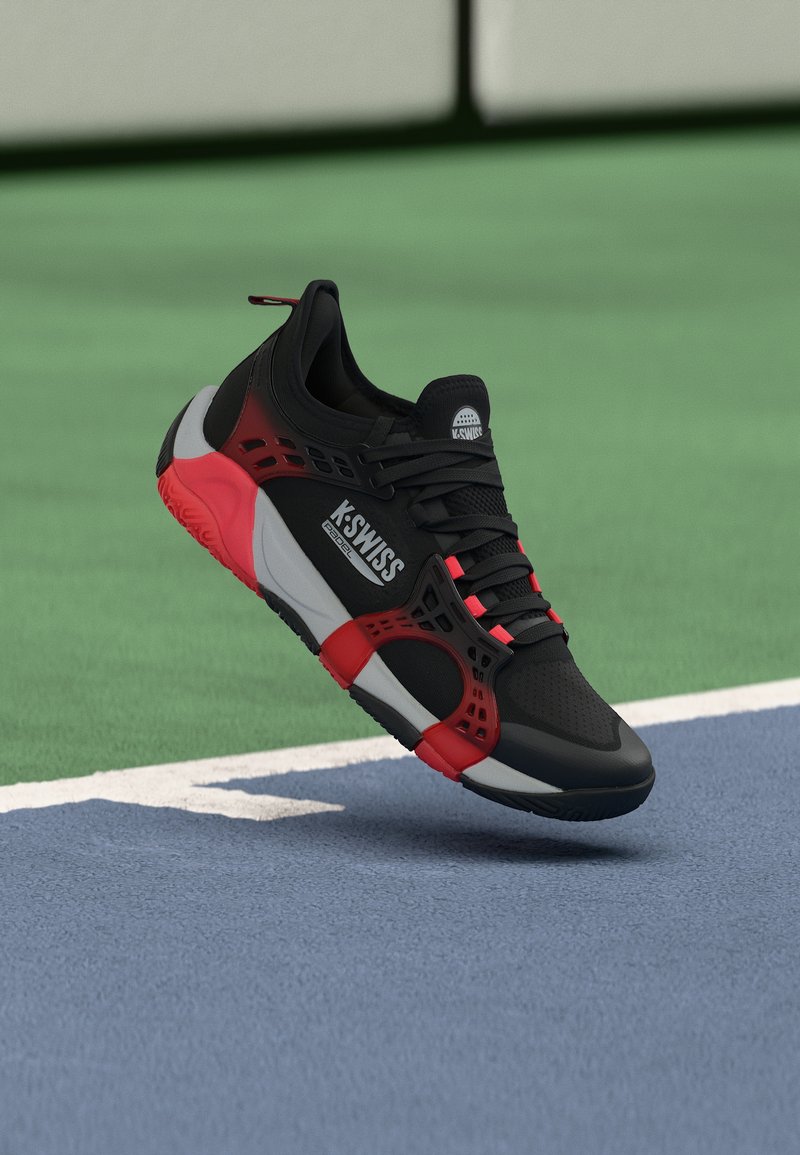 Μαύρο και κόκκινο αθλητικό παπούτσι K-Swiss με κορδόνια, να αιωρείται πάνω από μια μπλε και πράσινη επιφάνεια γηπέδου τένις.