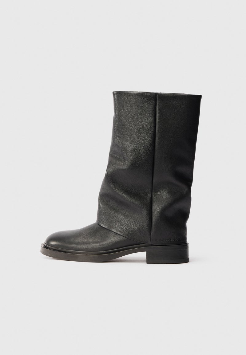 Μαύρη δερμάτινη μπότες μέχρι την κνήμη με χαμηλό τετράγωνο τακούνι και στρογγυλή μύτη, που εμφανίζει ελαφρές πτυχές και λεπτή υφή στην λεία επιφάνεια.