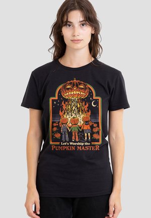 Frau mit schwarzem T-Shirt zeigt Halloween-Design mit drei Kindern, die Händchen halten, Kürbisköpfen, schwarzer Katze, Lagerfeuer und dem Text "Lasst uns den Kürbis-Meister anbeten".