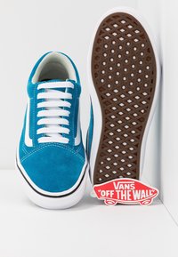 Modré semišové tenisky Vans s bílými tkaničkami a doplňky. Mají gumovou podrážku s diamantovým vzorem a červenou značkovou nášivkou.