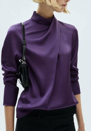 Une femme porte un chemisier violet à col montant avec des manches froncées et un petit sac à épaule noir sur un fond uni.