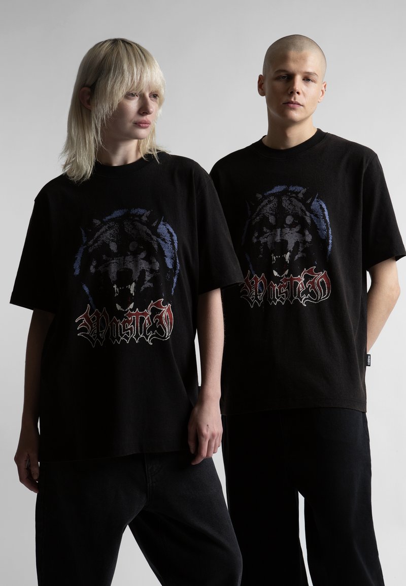 Δύο άτομα φορούν μαύρα, φαρδιά μπλουζάκια με τυπωμένο σκίτσο λύκου που γρυλίζει και το κείμενο «Wasted», στέκονται μπροστά σε απλό γκρι φόντο.