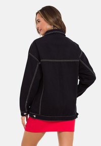 Schwarze Jeansjacke mit Kontrastnähten, Kragen und seitlichen Knopfdetails, getragen über einem roten gerippten Kleid.
