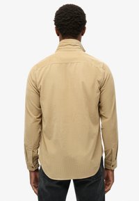 Mężczyzna z krótkimi kręconymi włosami, ubrany w beżową koszulę z długim rękawem i ciemne spodnie, stojący tyłem na tle białego tła.