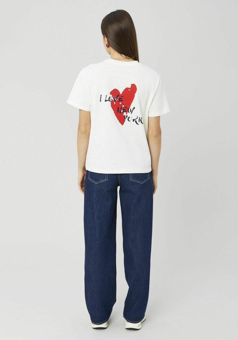 T-shirt en coton blanc arborant un graphique de grand cœur rouge vif avec le texte « J'AIME NEW YORK » en noir, associé à un jean en denim foncé à jambes larges.
