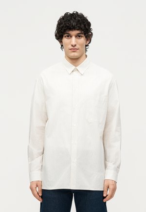 Jonge man met krullend haar die een wit overhemd met lange mouwen en subtiele verticale strepen draagt, staand tegen een effen achtergrond.