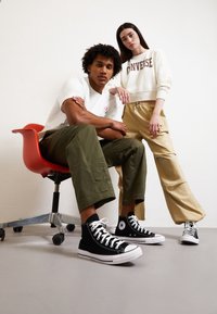 Czarne sneakersy za kostkę z białymi sznurówkami i gumowymi podeszwami, w połączeniu z oliwkowymi spodniami o szerokich nogawkach i kremową bluzą z napisem "Converse".