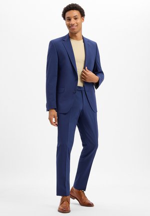 Man wearing a blue suit with beige shirt and brown dress shoes standing and smiling against a white background.
