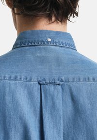 Camicia di jeans azzurro chiaro con yoke sul retro, bottone a pressione al colletto e una superficie liscia con una leggera lucentezza.