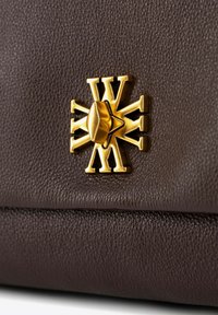 Bruine leren tas met een gestructureerd oppervlak, voorzien van een gouden geometrisch embleem met een centrale stenen accent, wat de ingewikkelde details en ambacht benadrukt.