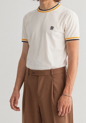 Witte t-shirt met contrasterende blauwe en gele geribde kraag en mouwen. Bevat een klein geborduurd logo op de borst. Gekozen in combinatie met een bruine broek.