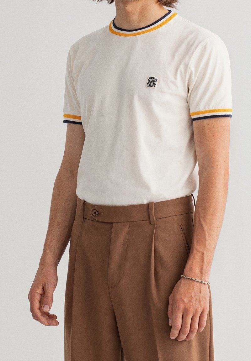 T-shirt blanc avec un col et des poignets côtelés bleu et jaune contrastants. Présente un petit logo brodé sur la poitrine. Porté avec un pantalon marron.