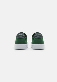Ténis verdes em lona com sola de borracha branca, apresentando um acabamento texturizado e um tabuleiro de calcanhar cinza impresso com "LACOSTE".